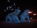 Two bear sculptures covered in blue lights are situated in an outdoor setting, illuminated against the dark night. Surrounding the sculptures are glowing streetlights, enhancing the night-time ambiance. The bears appear to be walking forward.