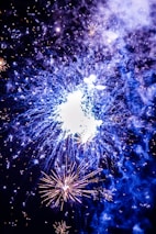 Bright fireworks illuminate the night sky with vivid bursts of light and color. The central explosion creates a striking display with surrounding smaller fireworks highlighting the scene. Smoke from the fireworks adds a textured effect, enhancing the overall spectacle.