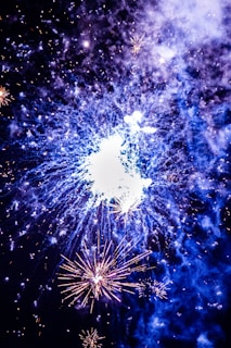 Bright fireworks illuminate the night sky with vivid bursts of light and color. The central explosion creates a striking display with surrounding smaller fireworks highlighting the scene. Smoke from the fireworks adds a textured effect, enhancing the overall spectacle.