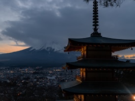 A traditional Japanese pagoda stands prominently in the foreground with intricate architectural details. In the background, Mount Fuji looms large, partially obscured by thick, dark clouds. The city below is dotted with lights as the sun sets, casting a soft orange and pink hue across the sky.