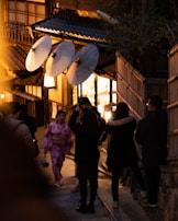 In a dimly lit, narrow street, a woman wearing a pink kimono is walking while holding a small purse. Traditional Japanese paper umbrellas are displayed on a building, creating a warm and inviting atmosphere. Several people dressed in dark winter clothing are taking photos or strolling along the street, which has wooden structures and ambient lighting.