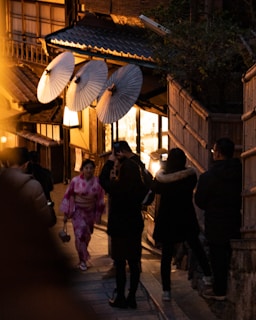 In a dimly lit, narrow street, a woman wearing a pink kimono is walking while holding a small purse. Traditional Japanese paper umbrellas are displayed on a building, creating a warm and inviting atmosphere. Several people dressed in dark winter clothing are taking photos or strolling along the street, which has wooden structures and ambient lighting.