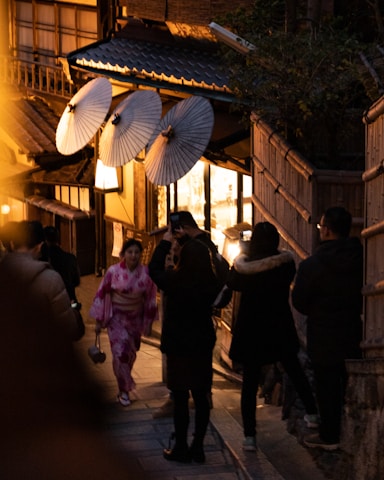 In a dimly lit, narrow street, a woman wearing a pink kimono is walking while holding a small purse. Traditional Japanese paper umbrellas are displayed on a building, creating a warm and inviting atmosphere. Several people dressed in dark winter clothing are taking photos or strolling along the street, which has wooden structures and ambient lighting.