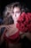 A woman poses dramatically against a dark background, wearing a red lingerie set with ruffled details. She has voluminous, wavy hair and striking makeup, including bold eye shadow. Her expression is intense, and she holds a large red flower that adds to the dramatic aesthetic.