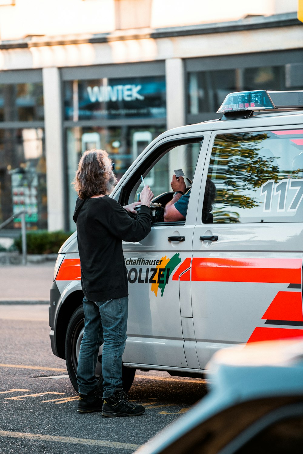 man in black jacket and blue denim jeans standing beside white and orange van during daytime