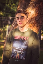 A young person stands outdoors near a tree, wearing a green sweatshirt with a graphic design featuring a car and text that reads 'HIGHWAY OUTLAWS'. The background includes a blurred fence and greenery, suggesting a park or garden setting.
