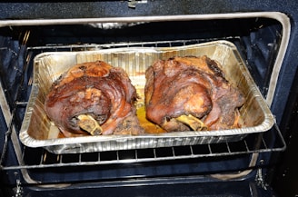 Two large, roasted pork knuckles situated in a foil tray inside an oven. The meat appears well-cooked, with a rich brown color and crispy exterior.