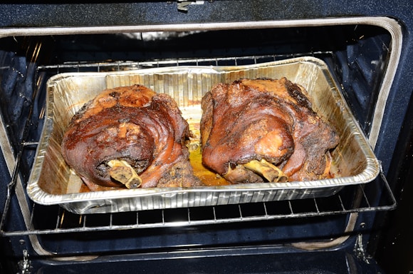 Two large, roasted pork knuckles situated in a foil tray inside an oven. The meat appears well-cooked, with a rich brown color and crispy exterior.