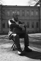 A bronze statue of a bearded man sits on a stool in front of an old building with large windows. The man appears contemplative, with his arms crossed over a bag resting on his lap. The statue is set in an outdoor area with bare trees, suggesting it might be early spring or late autumn.