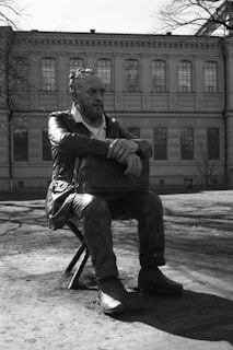 A bronze statue of a bearded man sits on a stool in front of an old building with large windows. The man appears contemplative, with his arms crossed over a bag resting on his lap. The statue is set in an outdoor area with bare trees, suggesting it might be early spring or late autumn.
