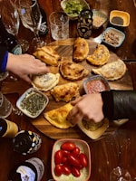 A wooden table holds a variety of food items centered around a serving platter filled with empanadas. Surrounding the platter are small bowls containing pesto, salsa, cherry tomatoes, herbs, and spices. Several hands are reaching for food, and there are bottles of beverages and glasses added to the lively table setting.