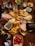 A wooden table holds a variety of food items centered around a serving platter filled with empanadas. Surrounding the platter are small bowls containing pesto, salsa, cherry tomatoes, herbs, and spices. Several hands are reaching for food, and there are bottles of beverages and glasses added to the lively table setting.