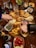 A wooden table holds a variety of food items centered around a serving platter filled with empanadas. Surrounding the platter are small bowls containing pesto, salsa, cherry tomatoes, herbs, and spices. Several hands are reaching for food, and there are bottles of beverages and glasses added to the lively table setting.