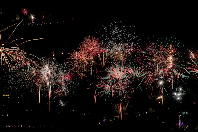 Vibrant fireworks burst in various shapes and sizes against the backdrop of a dark night sky. The display features vivid colors, primarily reds, greens, and whites, creating a dazzling spectacle.
