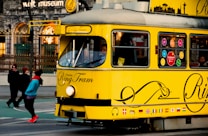 A vibrant yellow tram adorned with various flags and decorative designs travels through an urban setting. A pedestrian in bright clothing walks nearby, and there are lights and architecture in the background suggesting a city environment.