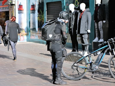 A person wearing motorcycle gear, including a helmet and protective clothing, stands beside a bicycle on a city street. The individual carries a backpack and appears to be inspecting or adjusting the bicycle. Nearby, there are mannequins in a shop display showcasing casual clothing. Other pedestrians walk along the street.