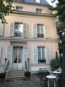 A charming building facade with light pink walls and white shutters. Three levels of windows have decorative wrought iron railings. A small patio in front features round white tables, black chairs, and potted plants. Large umbrellas and trees frame the relaxing outdoor space.