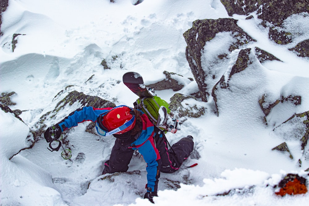 Hombre con chaqueta roja y pantalones azules sentado en el suelo cubierto de nieve durante el día