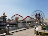 Amusement park scene featuring a large ferris wheel with a recognizable character's face, a roller coaster with red tracks, and a body of water in the foreground. The area is landscaped with palm trees and decorative lampposts lining the walkway.