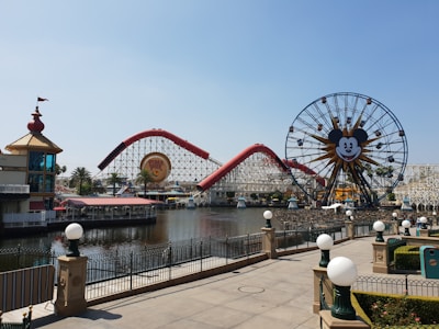 Amusement park scene featuring a large ferris wheel with a recognizable character's face, a roller coaster with red tracks, and a body of water in the foreground. The area is landscaped with palm trees and decorative lampposts lining the walkway.