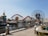 Amusement park scene featuring a large ferris wheel with a recognizable character's face, a roller coaster with red tracks, and a body of water in the foreground. The area is landscaped with palm trees and decorative lampposts lining the walkway.