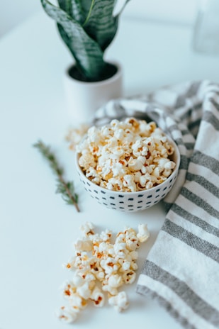 A bowl filled with popcorn placed on a white surface beside a gray and white striped cloth. A green potted plant is visible in the background, adding a touch of nature to the scene. A small sprig of greenery lies next to the popcorn, enhancing the fresh and natural look.
