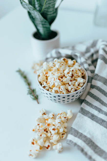 A bowl filled with popcorn placed on a white surface beside a gray and white striped cloth. A green potted plant is visible in the background, adding a touch of nature to the scene. A small sprig of greenery lies next to the popcorn, enhancing the fresh and natural look.