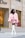 A woman is crossing a street wearing a stylish pink fur coat and a matching pink mini skirt. She accessorizes with white knee-high boots, white sunglasses, and carries a small pink handbag. A high-end fashion store is in the background.