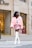 A woman is crossing a street wearing a stylish pink fur coat and a matching pink mini skirt. She accessorizes with white knee-high boots, white sunglasses, and carries a small pink handbag. A high-end fashion store is in the background.