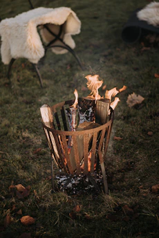 A metal fire pit with burning logs is situated on a grassy area. Nearby, a chair draped with a soft, cream-colored blanket provides a cozy atmosphere. Fallen leaves are scattered around the scene, enhancing the autumnal setting.