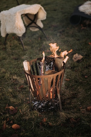 A metal fire pit with burning logs is situated on a grassy area. Nearby, a chair draped with a soft, cream-colored blanket provides a cozy atmosphere. Fallen leaves are scattered around the scene, enhancing the autumnal setting.