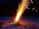 A tennis ball lies on a rough, textured surface, emitting a burst of bright, fiery sparks that shoot upward in a dynamic pattern. The background is dark, highlighting the vibrant glow of the flames and the texture of the ground.