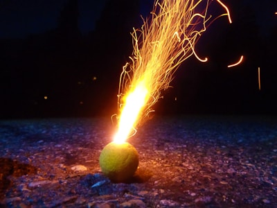 A tennis ball lies on a rough, textured surface, emitting a burst of bright, fiery sparks that shoot upward in a dynamic pattern. The background is dark, highlighting the vibrant glow of the flames and the texture of the ground.