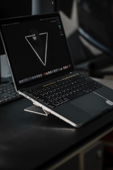 A sleek laptop placed on a dark surface with the screen displaying a minimalist wallpaper featuring a neon triangle with a wolf inside it. The laptop is partially open, and the keyboard keys are visible. The background is slightly blurred, hinting at an indoor setting.