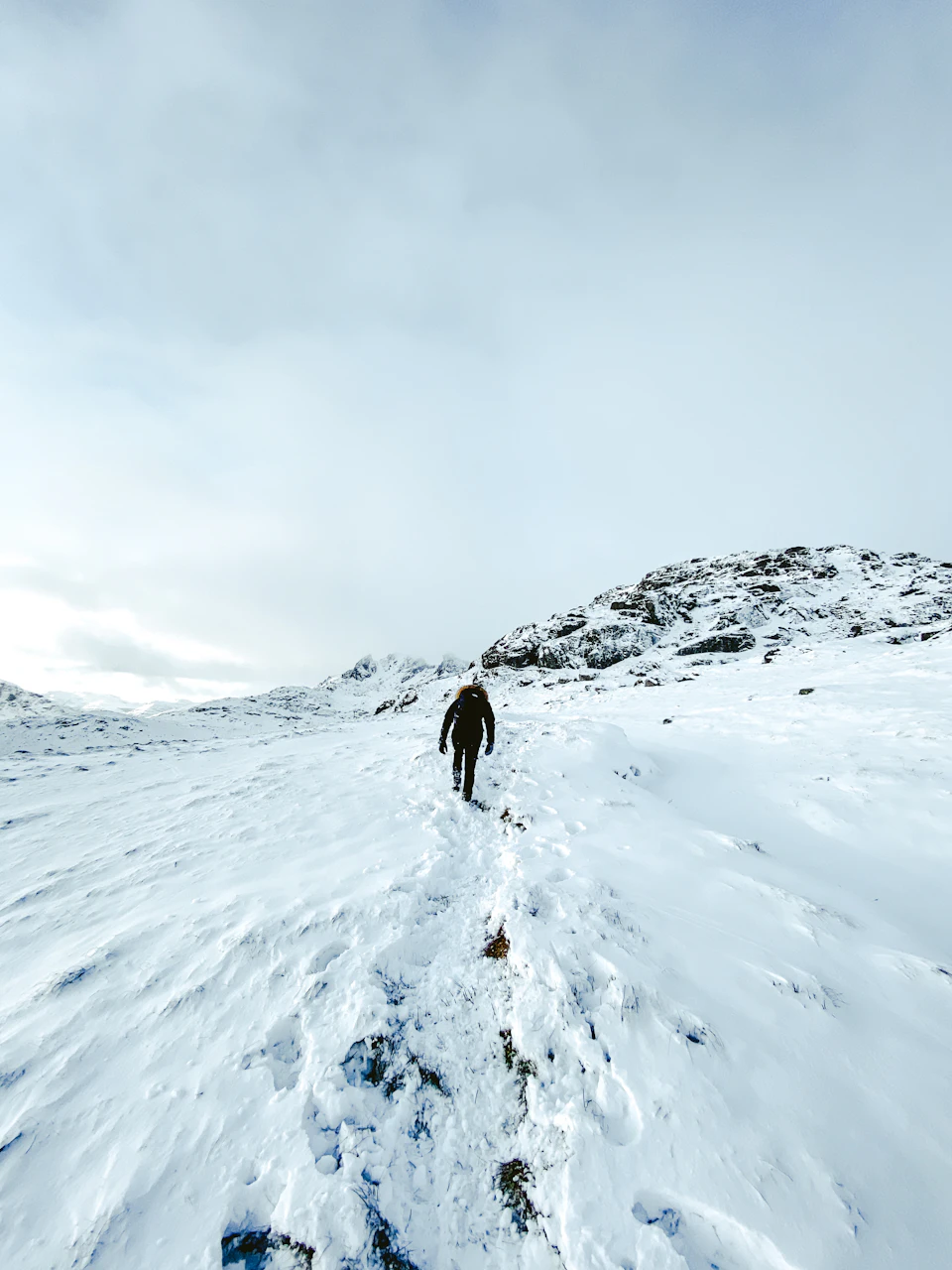 The image depicts a lone hiker walking away from the camera on a snowy mountain trail, surrounded by snow-covered rocks and trees. The sky above is overcast with clouds, casting a soft light over the scene.