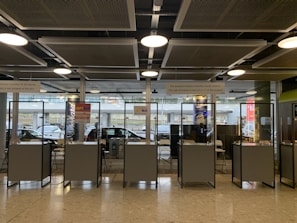 Several booth counters with protective glass are arranged in a row. Behind them, there are glass windows that provide a view of parked cars outside. Signs above the counters indicate services related to minibus transfers to ski resorts. The lighting is provided by ceiling lights and there is a modern industrial ceiling.