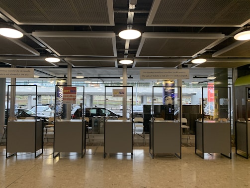 Several booth counters with protective glass are arranged in a row. Behind them, there are glass windows that provide a view of parked cars outside. Signs above the counters indicate services related to minibus transfers to ski resorts. The lighting is provided by ceiling lights and there is a modern industrial ceiling.