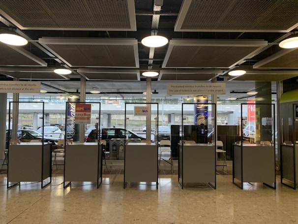 Several booth counters with protective glass are arranged in a row. Behind them, there are glass windows that provide a view of parked cars outside. Signs above the counters indicate services related to minibus transfers to ski resorts. The lighting is provided by ceiling lights and there is a modern industrial ceiling.