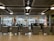 Several booth counters with protective glass are arranged in a row. Behind them, there are glass windows that provide a view of parked cars outside. Signs above the counters indicate services related to minibus transfers to ski resorts. The lighting is provided by ceiling lights and there is a modern industrial ceiling.