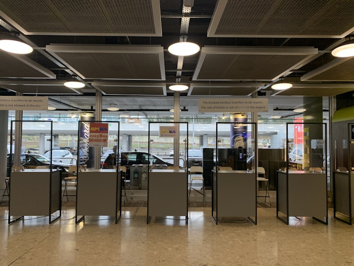 Several booth counters with protective glass are arranged in a row. Behind them, there are glass windows that provide a view of parked cars outside. Signs above the counters indicate services related to minibus transfers to ski resorts. The lighting is provided by ceiling lights and there is a modern industrial ceiling.