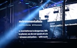 A dark stage setting is illuminated by bright beams of light and large LED screens displaying text related to environmentalists. The setup includes truss structures, lighting equipment, and hanging screens. The atmosphere suggests an event or concert with visual elements projecting environmental themes.