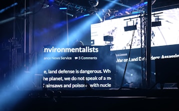 A dark stage setting is illuminated by bright beams of light and large LED screens displaying text related to environmentalists. The setup includes truss structures, lighting equipment, and hanging screens. The atmosphere suggests an event or concert with visual elements projecting environmental themes.