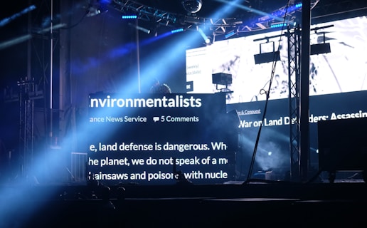 A dark stage setting is illuminated by bright beams of light and large LED screens displaying text related to environmentalists. The setup includes truss structures, lighting equipment, and hanging screens. The atmosphere suggests an event or concert with visual elements projecting environmental themes.
