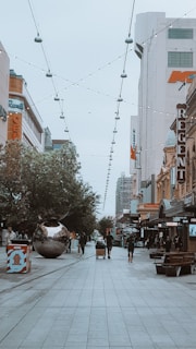 A lively urban street scene featuring a pedestrian walkway lined with shops and buildings. A large, reflective spherical sculpture is situated on the left, surrounded by trees. Pedestrians walk along the street, and string lights are suspended above.