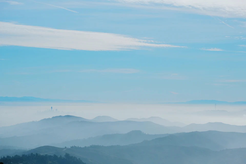 The image depicts a breathtaking view of a mountain range under a clear blue sky with wispy clouds. The mountains, painted in various shades of green and brown, stretch across the horizon, their peaks touching the sky at different elevations. The perspective is from below, looking up towards the majestic landscape. In the distance, there are several towers scattered throughout the scene, adding an element of intrigue to the natural beauty.