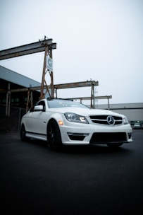 A sleek, white Mercedes-Benz car is parked on a dark asphalt surface. The environment consists of industrial structures with metal beams and a warehouse in the background, giving an urban and rugged feel.
