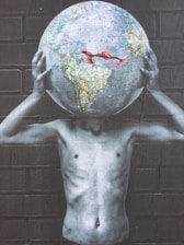 A surreal image depicting a shirtless figure holding a large globe that replaces their head against a brick wall backdrop. The depicted earth features clear details of continents and oceans, contrasted with grayscale arms and torso, creating a striking visual statement.
