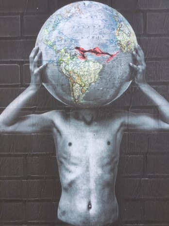 A surreal image depicting a shirtless figure holding a large globe that replaces their head against a brick wall backdrop. The depicted earth features clear details of continents and oceans, contrasted with grayscale arms and torso, creating a striking visual statement.