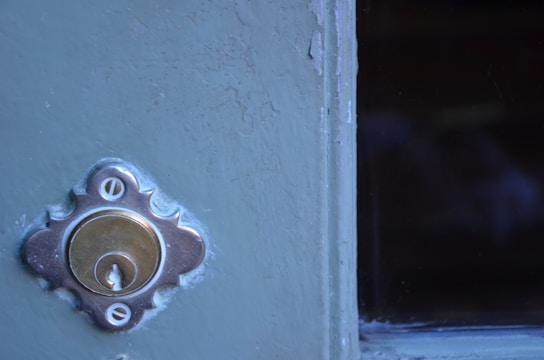 A close-up view of a metallic door lock set in a painted surface, with visible scratches and wear on the paint. To the right, there is a dark glass pane.