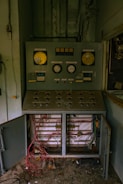 An old, industrial control panel is mounted against a wall, with dials, gauges, and various buttons. Below, tangled wires are exposed inside an open cabinet, suggesting disrepair or neglect. The surrounding area appears dusty and abandoned.